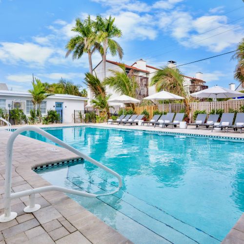 A sunny backyard pool with lounge chairs and white umbrellas, surrounded by palm trees and white buildings.