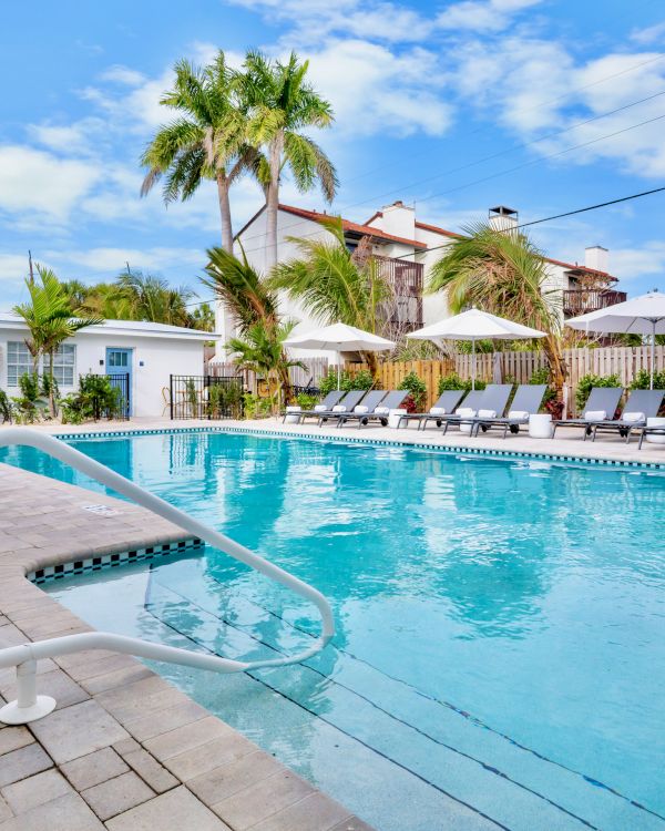 A sunny backyard pool with lounge chairs and white umbrellas, surrounded by palm trees and white buildings.