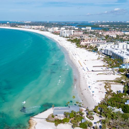Aerial view of a tropical beachfront with turquoise water, sand, hotels, and palm trees along the coastline.