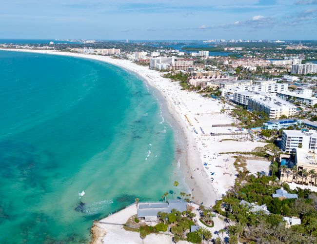 Aerial view of a tropical beachfront with turquoise water, sand, hotels, and palm trees along the coastline.