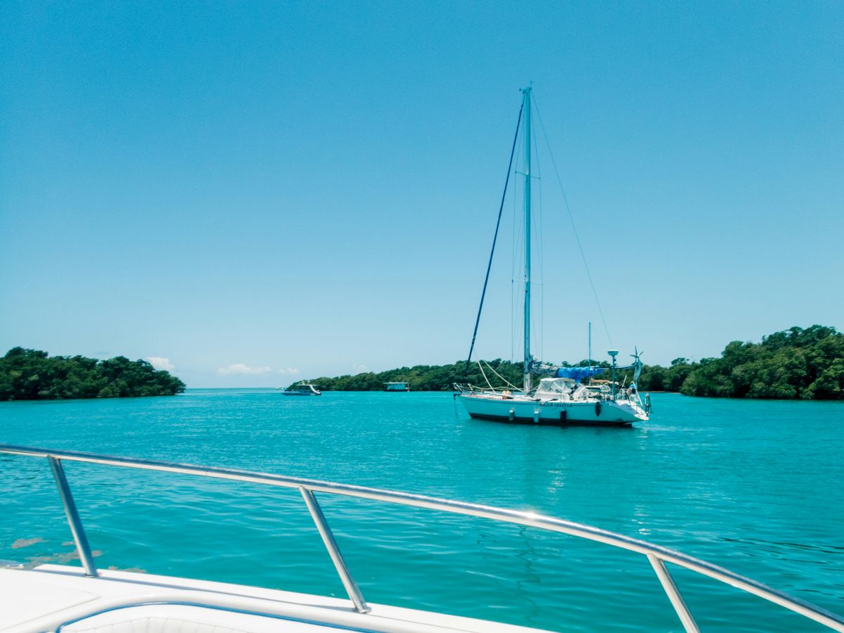 A sailboat with a tall mast sits on turquoise water near a green shoreline, seen from another boat under a bright blue sky.