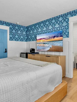 A bright modern bedroom with blue damask wallpaper, a mounted ocean sunset photo, a bed, and an attached vanity bath area.