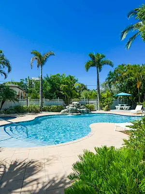 A sunny tropical pool with clear blue water, lounge chairs, palm trees, and a bright blue sky.