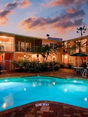 An outdoor hotel courtyard pool with palm trees and sun loungers, lit at dusk beneath colorful clouds.
