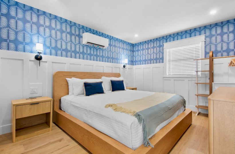 A modern bedroom with a wooden bed, blue leaf-pattern wallpaper, wall-mounted AC, bedside tables, and a window with blinds.