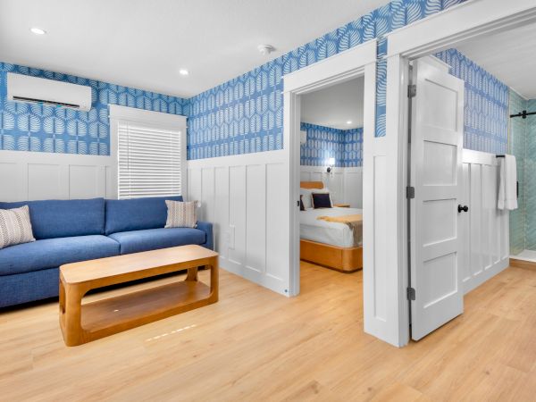 A bright living room with a blue sofa, wooden coffee table, patterned blue walls, and an open door revealing a bedroom and tiled bathroom.
