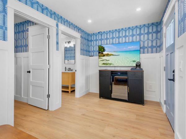 Bright modern room with blue leaf wallpaper, an open door to a sink, and a TV on a cabinet showing a beach scene.