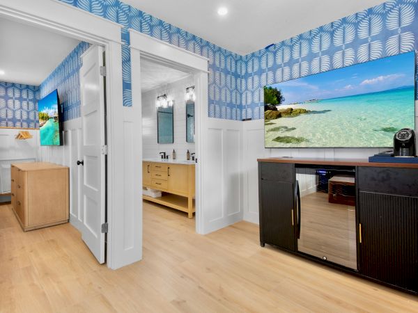 Bright modern room with blue leaf wallpaper, white vanity and sinks, two doors open, and a screen showing a turquoise beach scene.