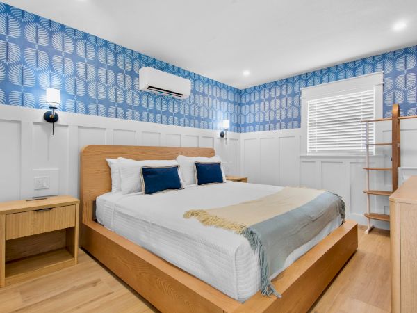 A modern bedroom with a wooden bed, blue feather-pattern wallpaper, two lamps, white walls, blinds, and a wall AC unit.