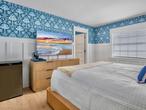 A modern bedroom with a bed, dresser, mounted TV, ocean-sunset wall art, blue floral wallpaper, and white blinds and AC.