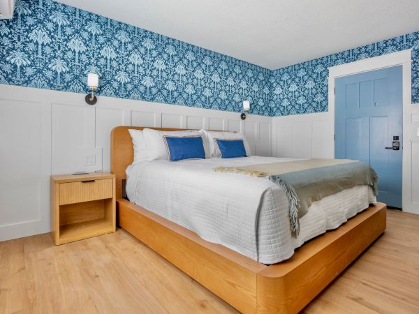 A bright bedroom with a large bed, blue patterned accent wall, two lamps, a wooden nightstand, and a blue door.