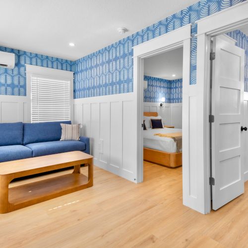 Bright room with a blue sofa and coffee table, blue leaf wallpaper, and a doorway to a tiled bathroom with a shower and towels.