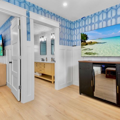 A bright modern bathroom/vanity area with blue leaf wallpaper, open doors, two wall-mounted screens, and a coffee machine on the counter.