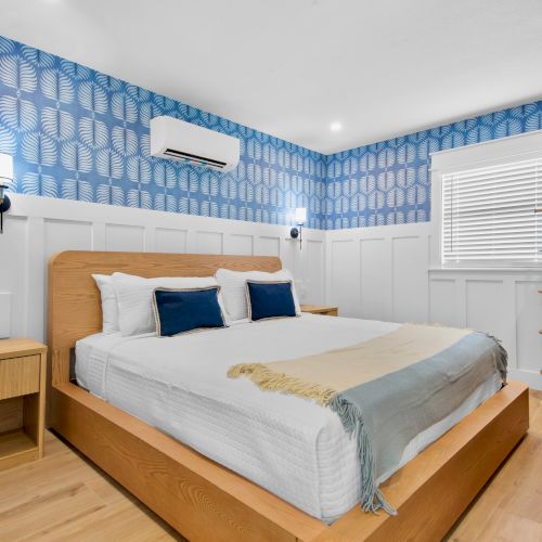 A modern bedroom with a wooden bed, blue/white patterned wallpaper, wall lights, air conditioner, and a window with blinds.