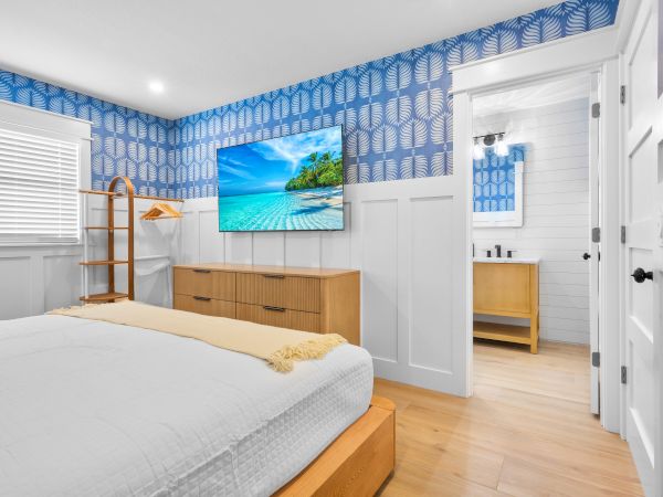 A bright modern bedroom with blue patterned wallpaper, a TV showing a tropical beach, and an open doorway to a clean bathroom.