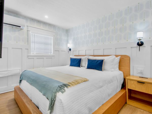 A bright bedroom with a white bed, blue pillows, wood headboard, wall lamps, and patterned wallpaper.