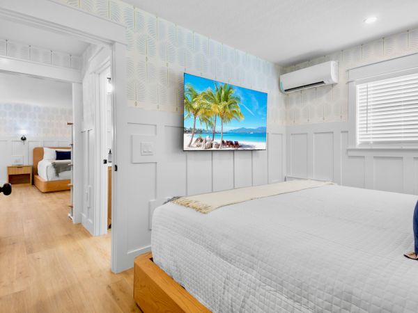 Bright modern bedroom with a bed, wall-mounted TV showing a beach scene, patterned wallpaper, and an air conditioner.
