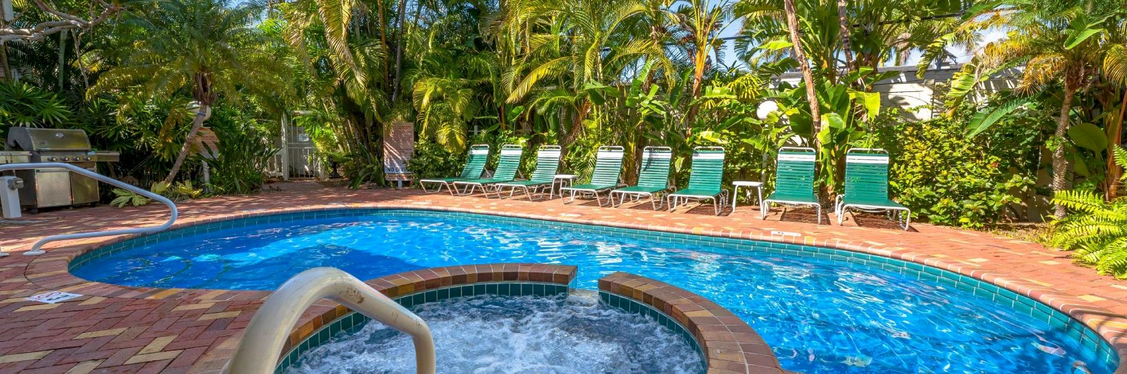 A bright outdoor swimming pool with a spa bubbler, surrounded by tropical palms and blue-green lounge chairs.