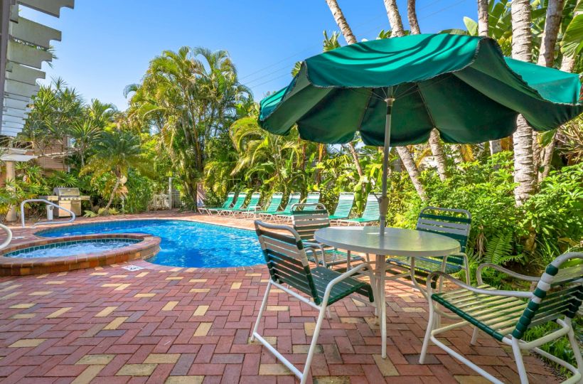 Tropical resort pool with lounge chairs, a green umbrella, palm trees, and a brick patio on a sunny day.