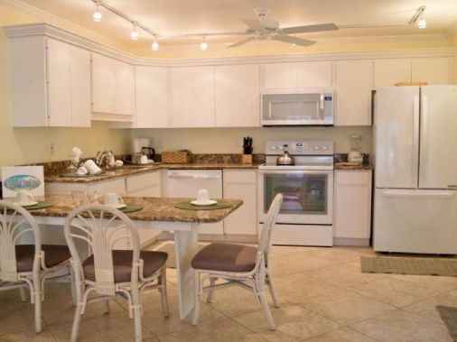 A bright modern kitchen with white cabinets, an oven and fridge, and a central island with several white dining chairs.