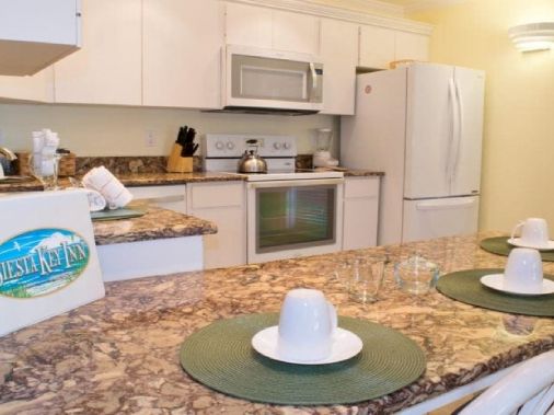 A bright hotel-style kitchen with granite countertops, a stove and microwave, a white refrigerator, and table settings with cups and plates.