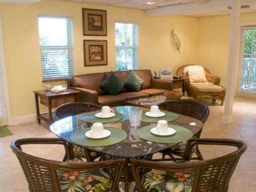 A bright living and dining room with wicker chairs, a round glass table, a sofa with pillows, and large sunlit windows.