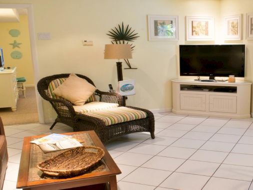 Bright living room with wicker chair, striped cushions, coffee table, framed wall art, and a flat-screen TV on a cabinet.