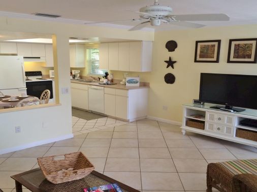 Bright living room with tiled floor, ceiling fan, TV on a white cabinet, and an open kitchen with light wood cabinets and countertops.