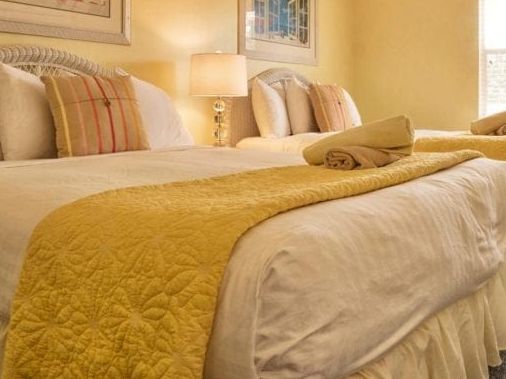 A neatly made hotel bedroom with soft yellow quilts, neatly arranged pillows, and bedside lamps.