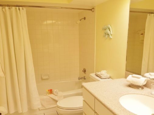 A hotel bathroom showing a bathtub with shower curtain, toilet, and a sink vanity with mirror and towels.