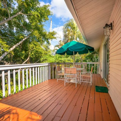 A cozy house porch with a wooden floor, two chairs, a green umbrella, and lush tropical trees in the background.