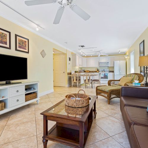 A bright living room and kitchen area with tiled floors, a flat-screen TV, sofas, and a ceiling fan.