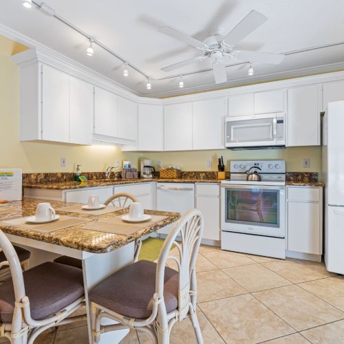 Bright modern kitchen with white cabinets, granite island, dining chairs, stove/oven, microwave, and fridge.