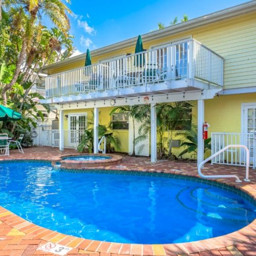 A bright resort courtyard with a blue swimming pool, patio loungers, palm trees, and a two-story building.