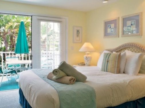 Sunlit bedroom with a large bed, pillows/comforter, bedside lamp, and French doors opening to a pool.