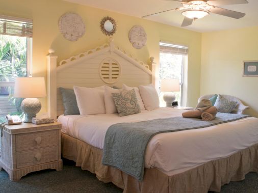 A bright cozy bedroom with a large white bed, patterned pillows, bedside tables, curtains, and framed wall art.