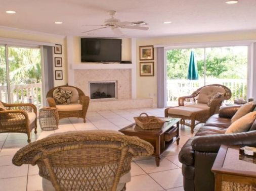 A bright living room with wicker chairs, leather sofa, coffee table, fireplace, TV, and large windows opening to lush greenery.