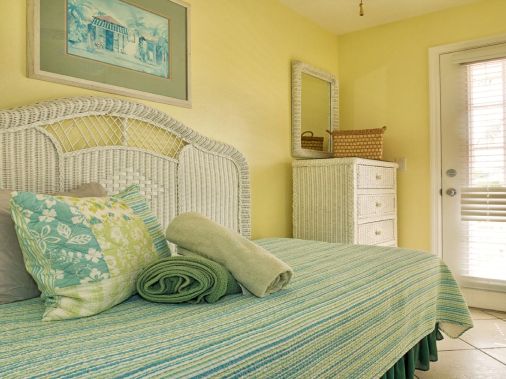 A cozy bedroom with a wicker bed, rolled blankets and pillows, and a dresser beside a bright window.