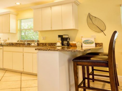 A bright modern kitchen with white cabinets, granite counters, a coffee maker, bar stools, and sunlit windows.
