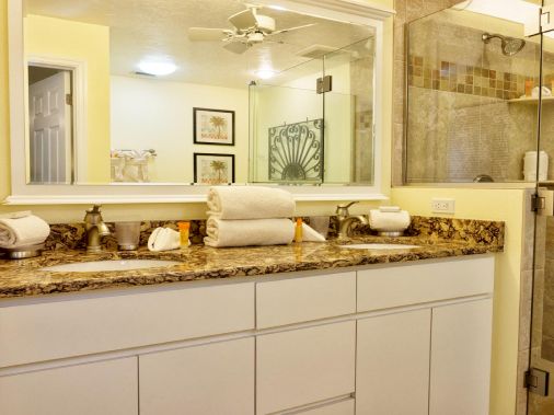 A bright bathroom with a granite vanity countertop, mirror above, and a glass-enclosed shower with tiled walls.