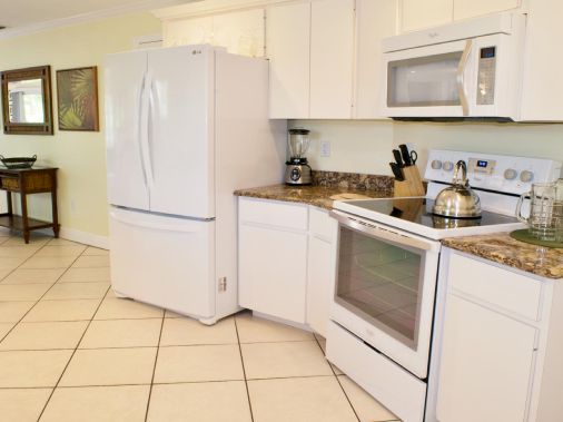 A bright white kitchen featuring a refrigerator, microwave, oven, countertop appliances, and tiled floors.