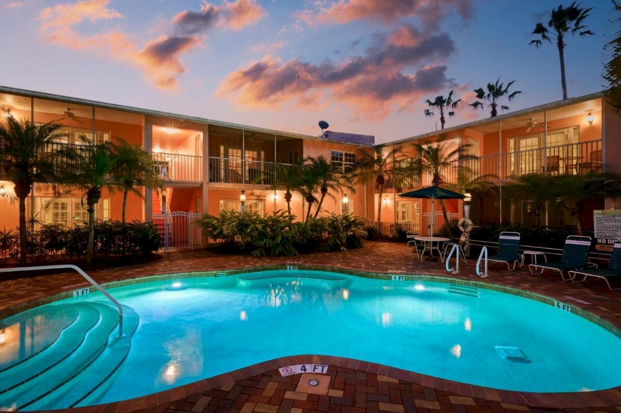 A hotel courtyard pool glows at dusk with sun loungers, palm trees, and two-story orange buildings under pink clouds.