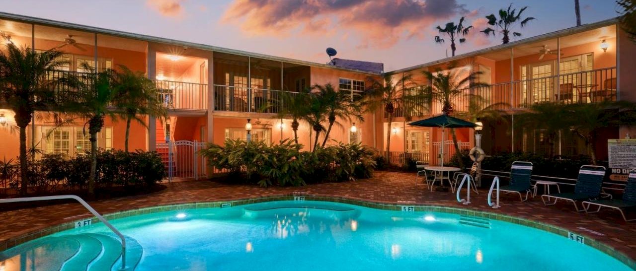 A hotel courtyard pool glows at dusk with sun loungers, palm trees, and two-story orange buildings under pink clouds.