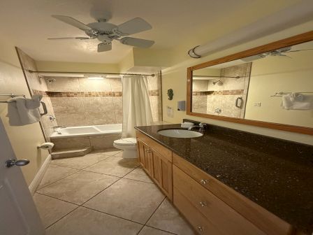 A bright bathroom with a tiled shower-tub, sink vanity, wall mirror, and ceiling fans with towels hanging.