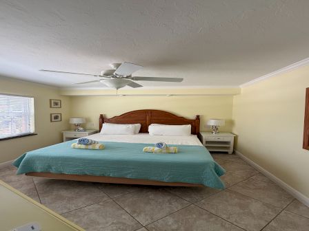 A bright bedroom with a wooden bed, white pillows, bedside tables, ceiling fan, and tiled floor.