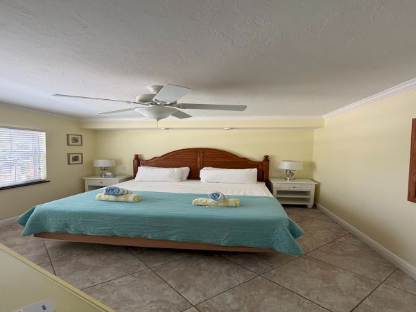 A bright bedroom with a wooden bed, white pillows, bedside tables, ceiling fan, and tiled floor.