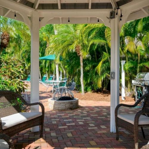 A sunny tropical courtyard patio with wicker lounge chairs, hanging lights, and lush palm trees around a fire-pit area.