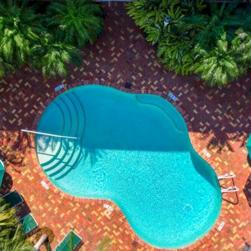Aerial view of a turquoise swimming pool surrounded by a brick patio, palm trees, lounge chairs, and a green umbrella.