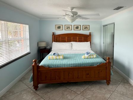 A tidy bedroom with a wooden bed, folded towels, ceiling fan, and windows with blinds plus framed wall art.