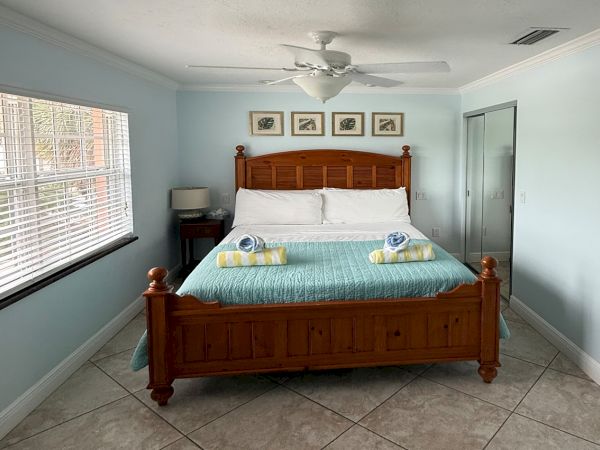 A tidy bedroom with a wooden bed, folded towels, ceiling fan, and windows with blinds plus framed wall art.